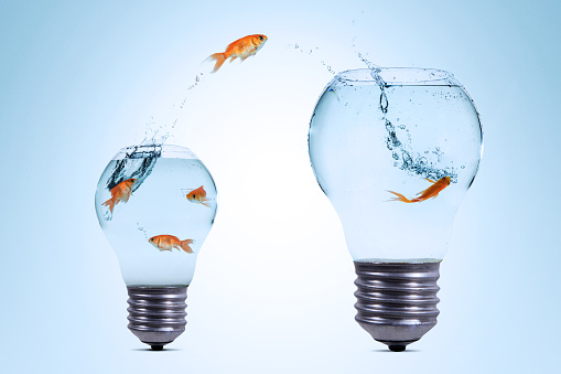 Gold fish jumping out from a smaller light bulb aquarium to a bigger one against blue background