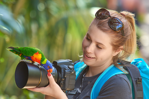 tourist smiling woman in tropical day holding camera, colorful parrot on it.