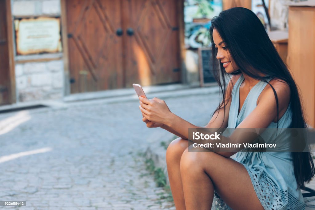 Young woman using smart phone on street Beautiful long haired girl sitting on ground and using smart phone, on street. Emoticon Stock Photo