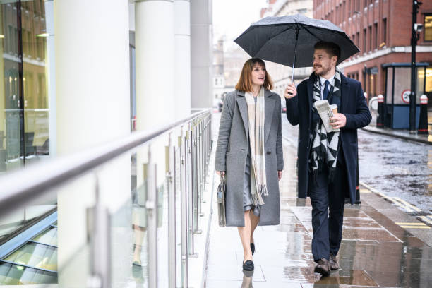 Business colleagues on way to walk under umbrella in the raing Man and woman in their 30s walking on wet pavement man, holding umbrella above their heads, woman talking chivalry stock pictures, royalty-free photos & images