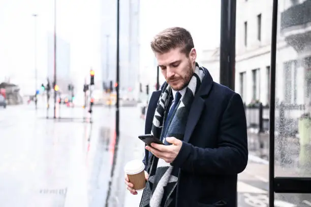 Photo of Mid adult businessman checking phone in bus stop with coffee