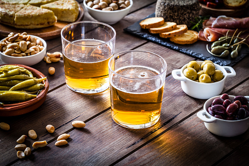 Two beer glasses shot on rustic wooden table with some tapas and appetizer. The beer glasses are at the center of an horizontal frame while some tapas are around them. The composition includes spanish tortilla, pickles, cheese, chorizo, bread, peanuts, pistachios, salami, prosciutto, jalapeño peppers, anchovies and others. DSRL studio photo taken with Canon EOS 5D Mk II and Canon EF 100mm f/2.8L Macro IS USM