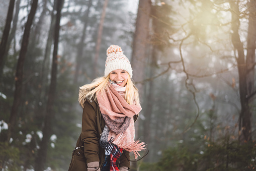 Young blonde women/girl in the forest. Surrounded with white snow. Girl is dressed in casual clothing, she is wearing olive coat, pastel pink scarf and hat, jeans and a red shirt. She is excited about snow, so she decided to go out on a walk, she is playing with snow and goofing around. Her big smile tells us how much she enjoys snow. Nature looks magical under the blanket of snow.
