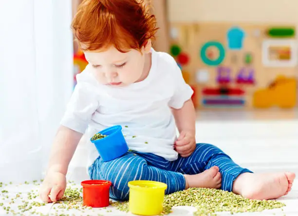 cute redhead baby boy developing his fine motility skills by playing with green peas at home