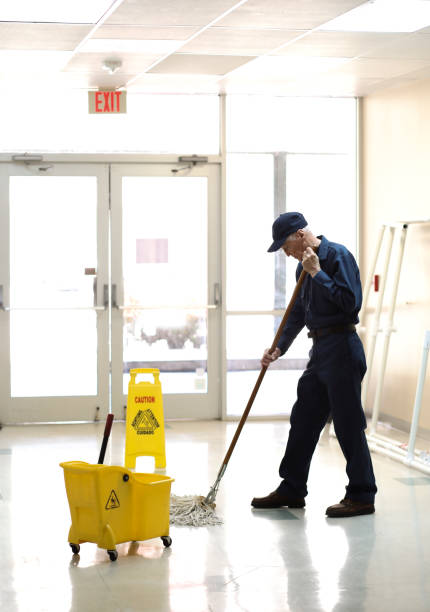 Here is a senior adult man working as a janitor in an office building, school or church.  He wears a uniform as he mops the entry floor.  Mop bucket and safety sign are also pictured.  He has retired and enjoys being active and still meeting people without responsibilities of the busy office demands.  Full length.