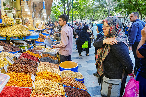 Tehran, Iran - April 29, 2017: A shopkeeper selling dried fruit in the Tehran Bazaar and elderly muslim woman dressed in hijab standing near him.