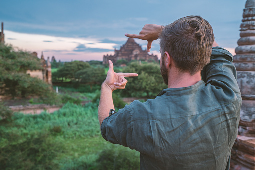 Caucasian young man contemplating the Bagan archeological zone at sunrise, temples pagodas and monastery coming out from the lush greenery. People travel concept.