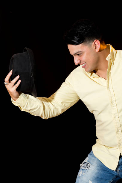 Young latin man poses in informal wear for a portrait. He is wearing a yellow shirt, jeans and a black hat. He is smiling and with his hat off. The background is black. The side flashlight has yellow gel.