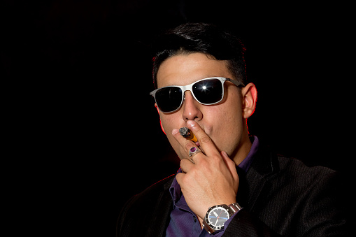 Young latin man poses in casual wear for a portrait. He is wearing a black jacket and blue shirt. He is smoking a cigar. The background is black. He is looking at camera.