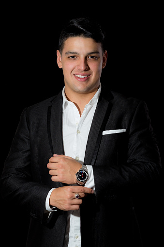 Young latin man poses in semi-formal wear for a portrait. He is wearing a black jacket, handkerchief on front pocket  and white shirt. He is serious. The background is black. He is fixing his sleeve.