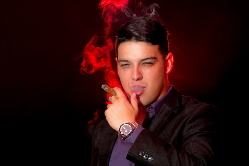 Young latin man poses in casual wear for a portrait. He is wearing a black jacket and blue shirt. He is smoking a cigar. The background is black. A red flashlight illuminates the smoke. He is looking at camera.
