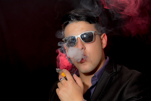 Young latin man poses in casual wear for a portrait. He is wearing a black jacket and blue shirt. He is smoking a cigar. The background is black. He is looking at camera. A red flashlight illuminates the smoke.