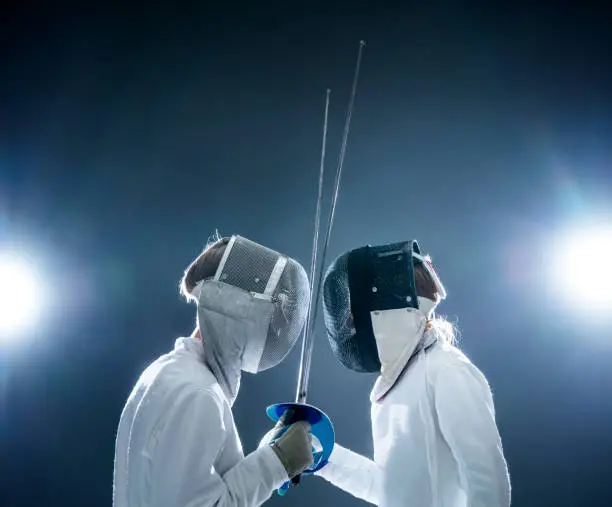 Side view of male fencers with foils standing face to face. Boys are wearing fencing costumes. Siblings are against illuminated black background.