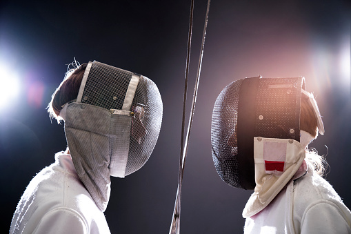 Side view of confident male fencers with foils standing face to face. Boys are wearing fencing costumes. Siblings are fencing against illuminated black background.