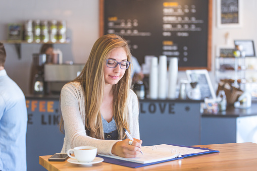 An attractive brunette college student studies in a modern coffee shop. She has her phone and coffee next to her and is writing notes in her binder. A barista is working in the background and there is a group at the table next to her.