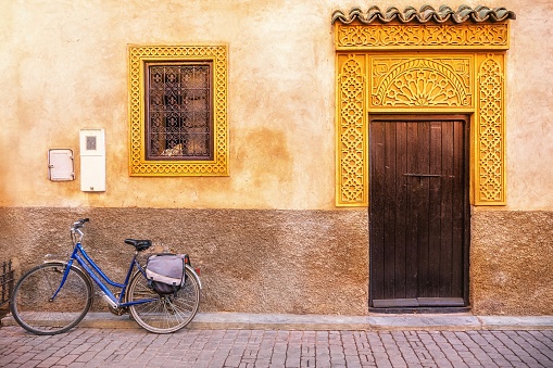 Very plain and simple house facade with some very elaborate gold colored window and door trim and grilles. A bicycle parked in front, on the cobblestone street.