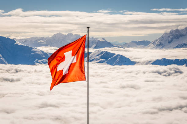 The red and white flag of Switzerland above a cloud filled valley in the alps with snow and glacier covered peaks in the background.