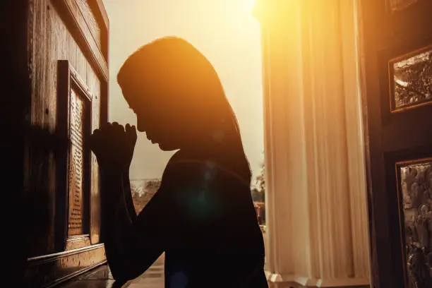 Photo of silhouette of woman kneeling and praying in modern church at sunset time