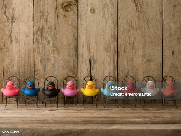 Line Of Of Many Different Colored Rubber Ducks Sitting On Chairs Facing Towards The Camera Scene Set In Front Of Old Wooden Paneling Stock Photo - Download Image Now