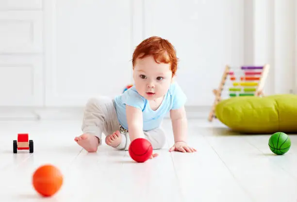 Photo of cute infant baby crawling on the floor at home, playing with colorful balls