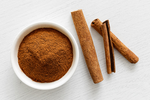 Finely ground cinnamon in white ceramic bowl isolated on white wood background from above. Cinnamon sticks.
