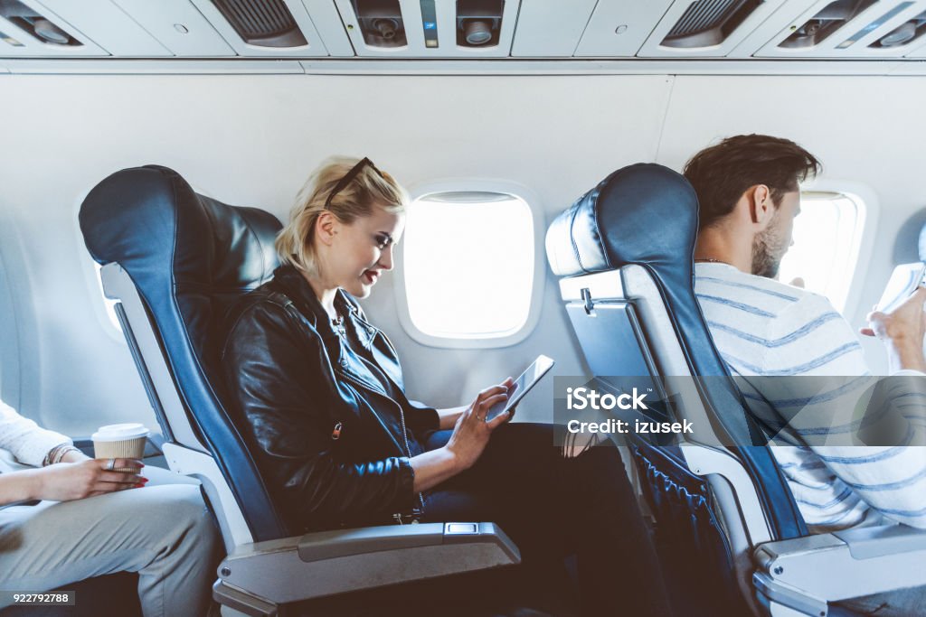 Female passenger using digital tablet during flight Young blonde woman sitting inside an airplane and using a digital tablet. Female passenger using e-reader during flight. Adult Stock Photo