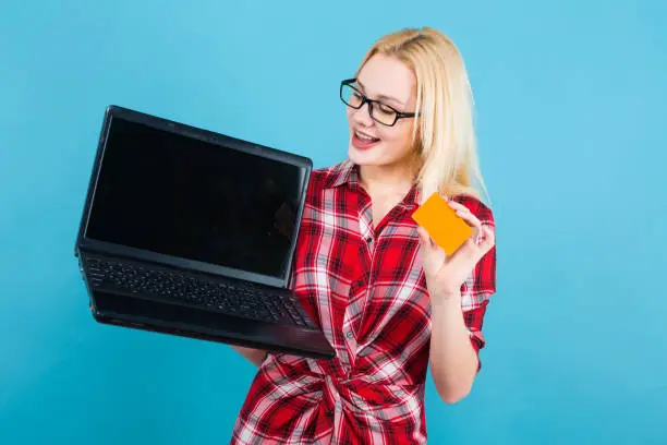 Portrait of attractive smiling blonde woman with glasses in red checkered shirt isolated on blue background holding laptop with blank business or credit card advertising loan concept