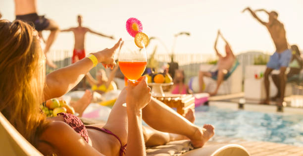 Woman lying by pool with cocktail in her hand while other people dance in and around pool at sunset Woman lying by pool with cocktail in her hand while her friends are dancing in and around pool at pool party at sunset. swimwear bikini top bikini bikini bottom stock pictures, royalty-free photos & images