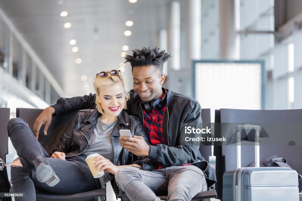 Young funky couple using mobile phone in airport lounge Multi ethnic young couple sitting on chair with suitcase in airport lounge and using mobile phone together. Adult Stock Photo