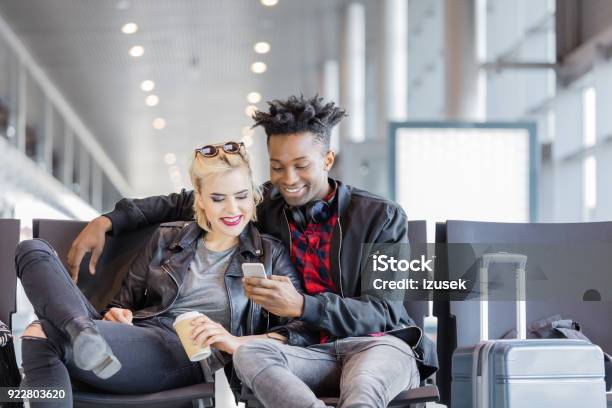Young Funky Couple Using Mobile Phone In Airport Lounge Stock Photo - Download Image Now