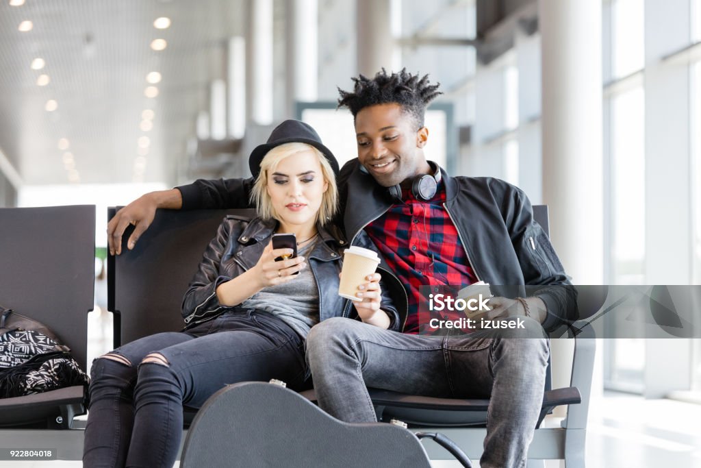 Young funky couple waiting for flight in airport lounge Multi ethnic young couple sitting on chair with suitcase in airport lounge and using  Airport Stock Photo