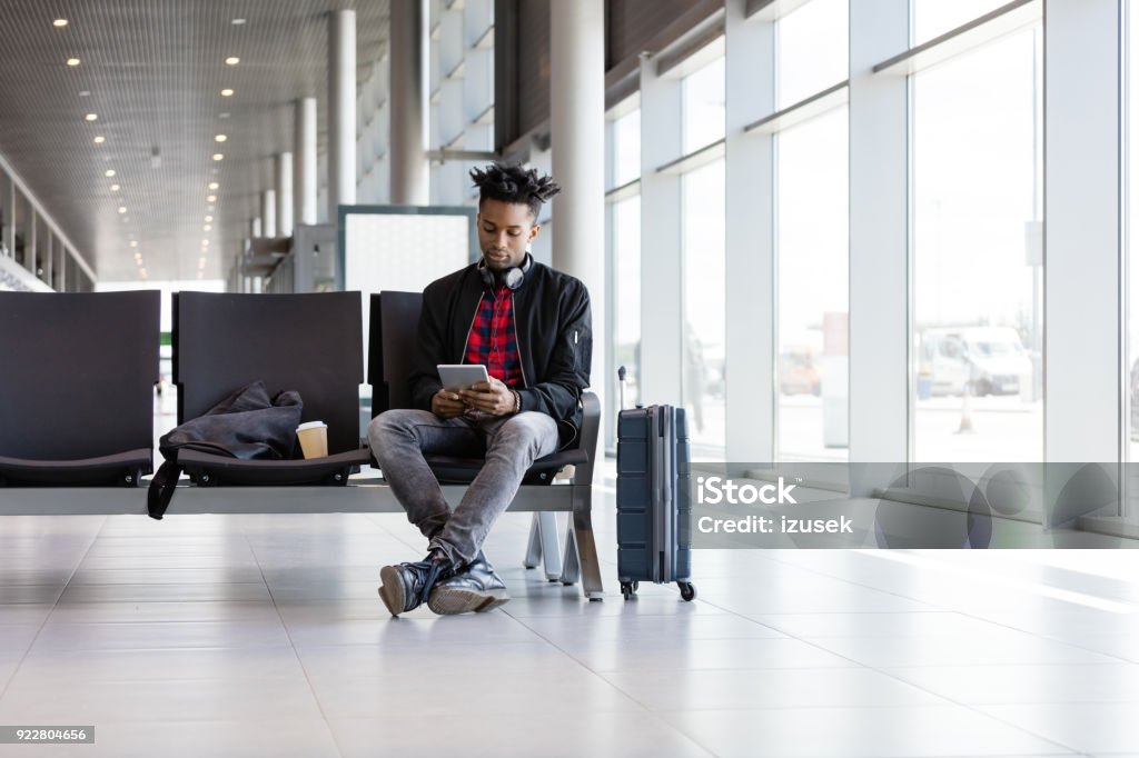 Young african using digital tablet in airport lounge Young African man sitting on chair with suitcase in airport lounge and using digital tablet. Men Stock Photo