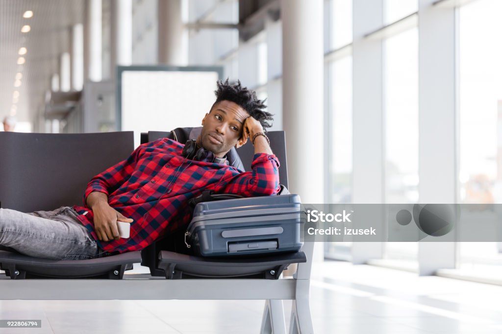 Young man waiting for flight in airport lounge Young african man lying on bench with suitcase in airport lounge, waiting for flight. Adult Stock Photo