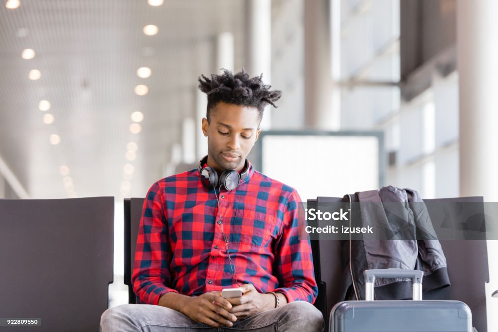 Young african using mobile phone in airport lounge Young african man sitting on chair with suitcase in airport lounge and using mobile phone. Adult Stock Photo