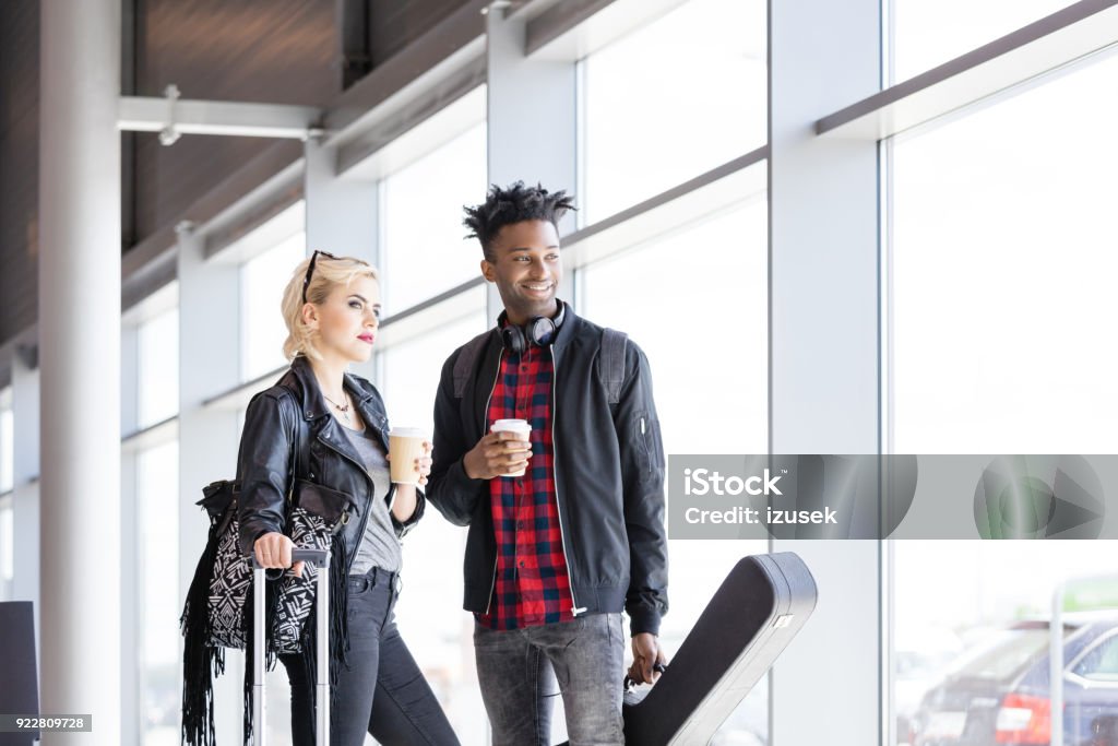 Young funky couple waiting for flight in airport lounge Multi ethnic young waiting for flight in airport lounge, drinking take away coffee. Young man holding guitar. Coffee - Drink Stock Photo