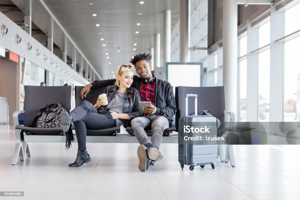 Multi ethnic funky couple waiting for flight in airport lounge Multi ethnic young couple sitting on chair with suitcase in airport lounge and using  Adult Stock Photo