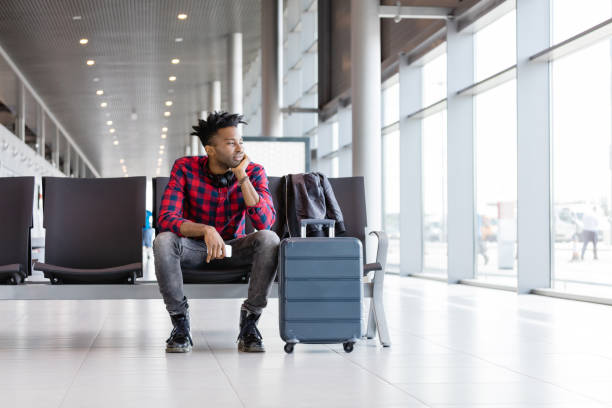 Young african man sitting on chair with suitcase in airport lounge, waiting for flight. Wide angle view.