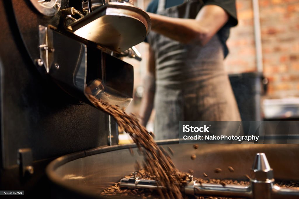 coffee beans being poured out of a coffee roasting machine African man wearing an apron busy pouring coffee beans from the coffee roasting machine over to the tray that stirs the beans until they are cool enough to be packaged. Coffee Roaster Stock Photo