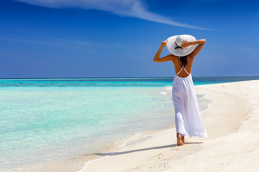 Happy traveler woman in white dress walks on a tropical beach