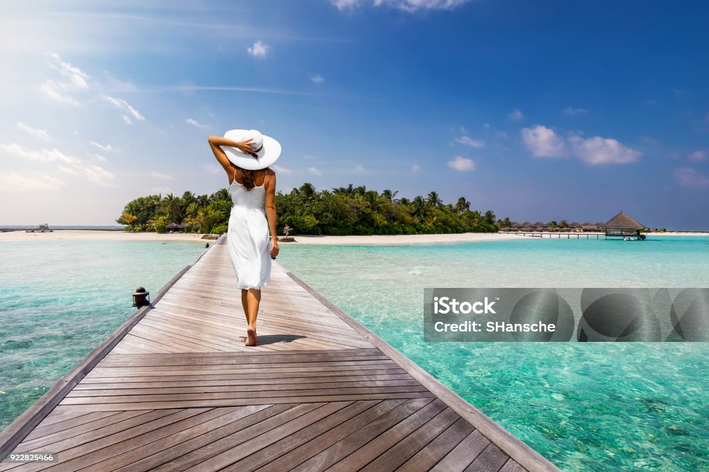 Attractive woman walks over a wooden jetty towards a tropical island Attractive woman in white walks over a wooden jetty towards a tropical island Luxury Stock Photo