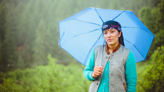 Asian, Indian beautiful traditional young woman enjoying fresh air in nature. She is looking at camera and standing under the umbrella by holding it in rainy weather. She is in traditional dress of Himachal Pradesh (Dathu and Askat). Shoot location Chopal, Himachal Pradesh.