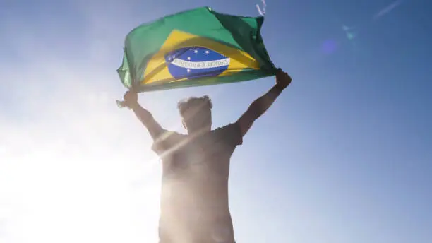 Young man holding brazilian national flag to the sky with two hands at the beach at sunset