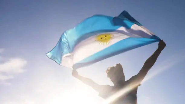 Photo of Young man holding argentinian national flag to the sky with two hands at the beach at sunset argentina