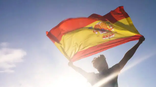 Photo of Young man holding spanish national flag to the sky with two hands at the beach at sunset spain