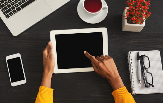 Black female hands holding tablet with blank screen for advertisement. Top view of african-american hands, laptop, smartphone and coffee on dark wooden table background, copy space