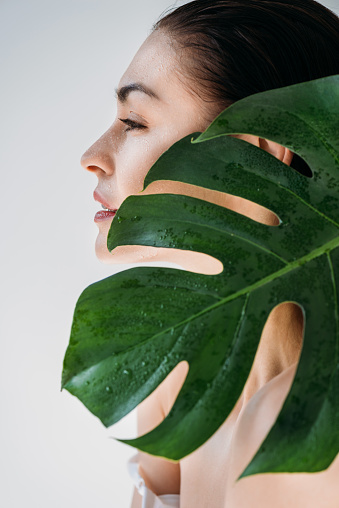 Side shot of young beautiful woman with wet fresh skin standing behind a green leaf