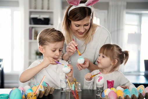 Mother with her daughter and son painting easter eggs at table. Family preparing for Easter. They are happy and positive, enjoying their time together, and proudly showing the colorful eggs. The sweet little girl is 2 years old and the boy is 4 years old. Easter holiday concept.