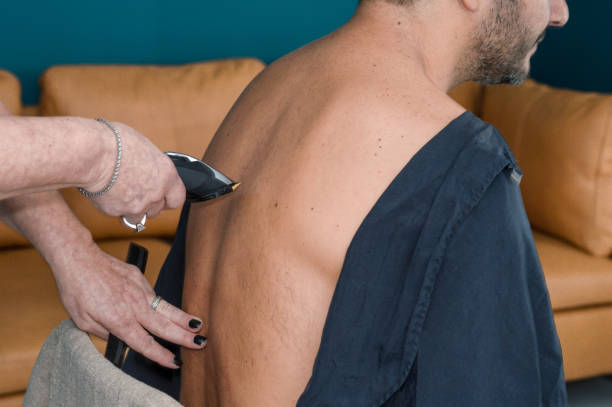 Female barber working with hair clipper, shaving young man's back stock photo