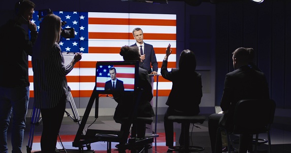 Official press conference of American representative politician on stage against display with American flag giving speech to audience in semilit studio and answers the questions.