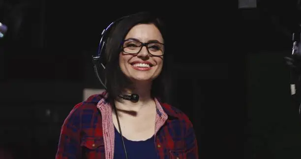 Casual young brunette in headset working in telecasting studio as stage director supervising footage making and cheerfully talking to colleagues.